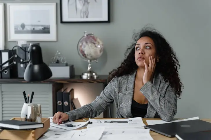 A woman wearing a plaid blazer sits at a table bored at work with her hand on her face.